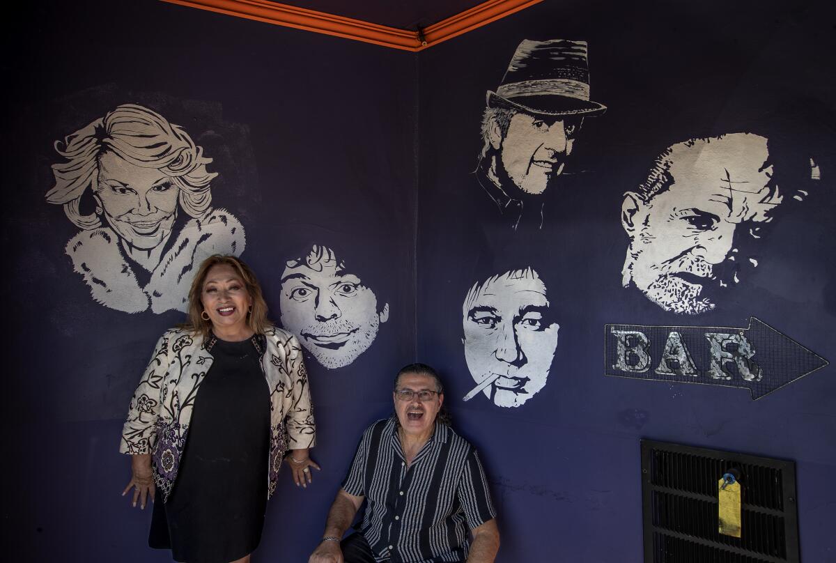 A man and woman leaning against a wall with painted faces of comedians on it