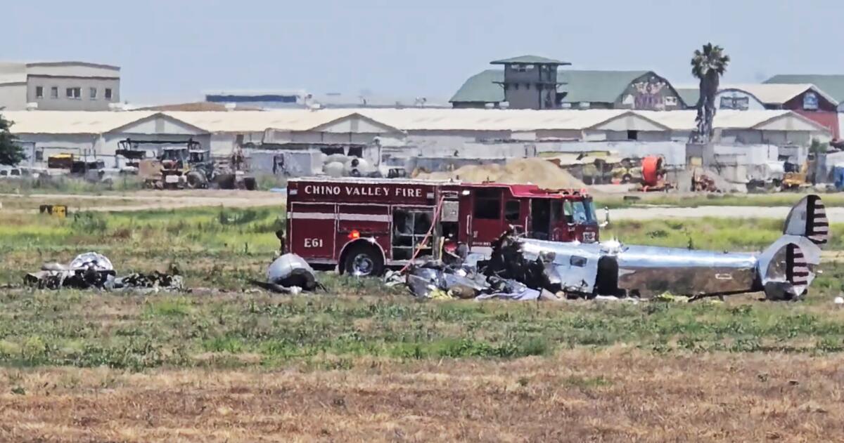 The wreckage of a twin-engine plane rests beside a fire engine in a field, with airport hangars in the background