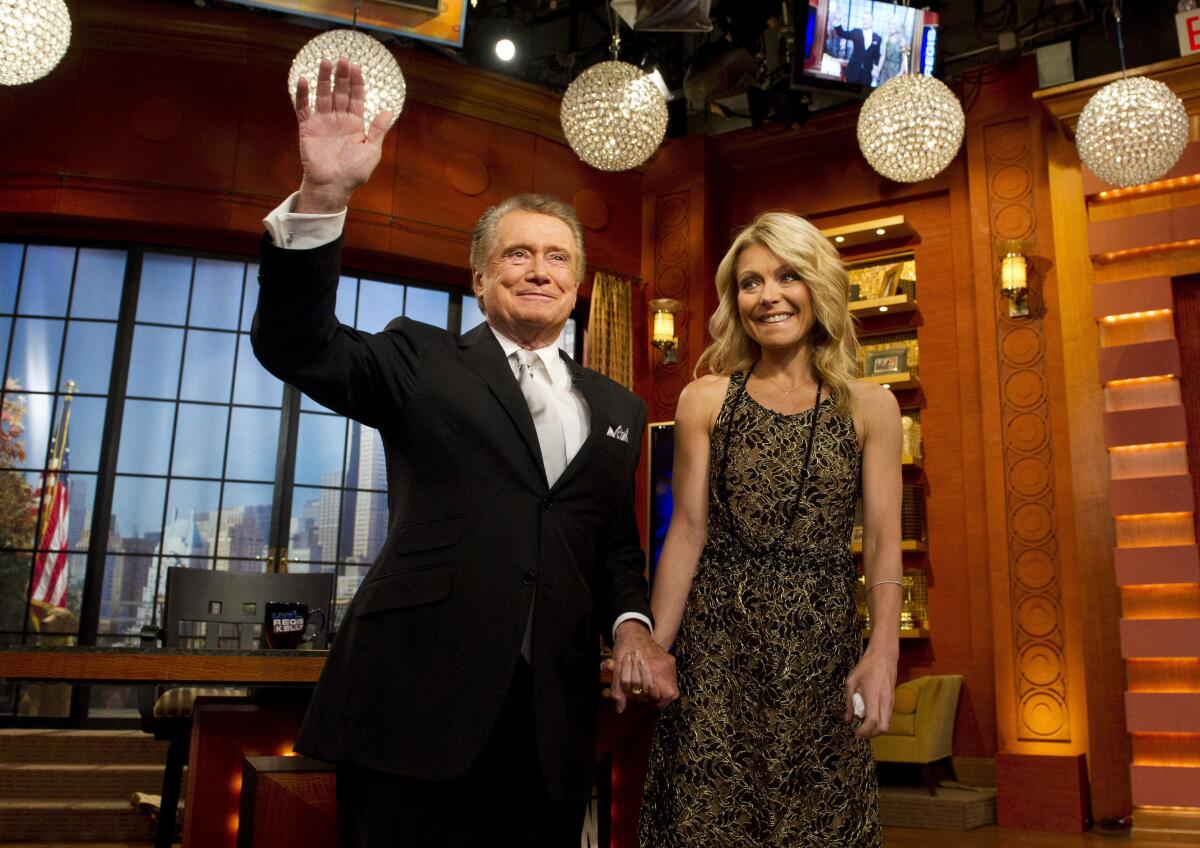 Man with gray hair wearing a suit and tie salutes with one hand while holding the hand of a blond woman wearing a brown dress