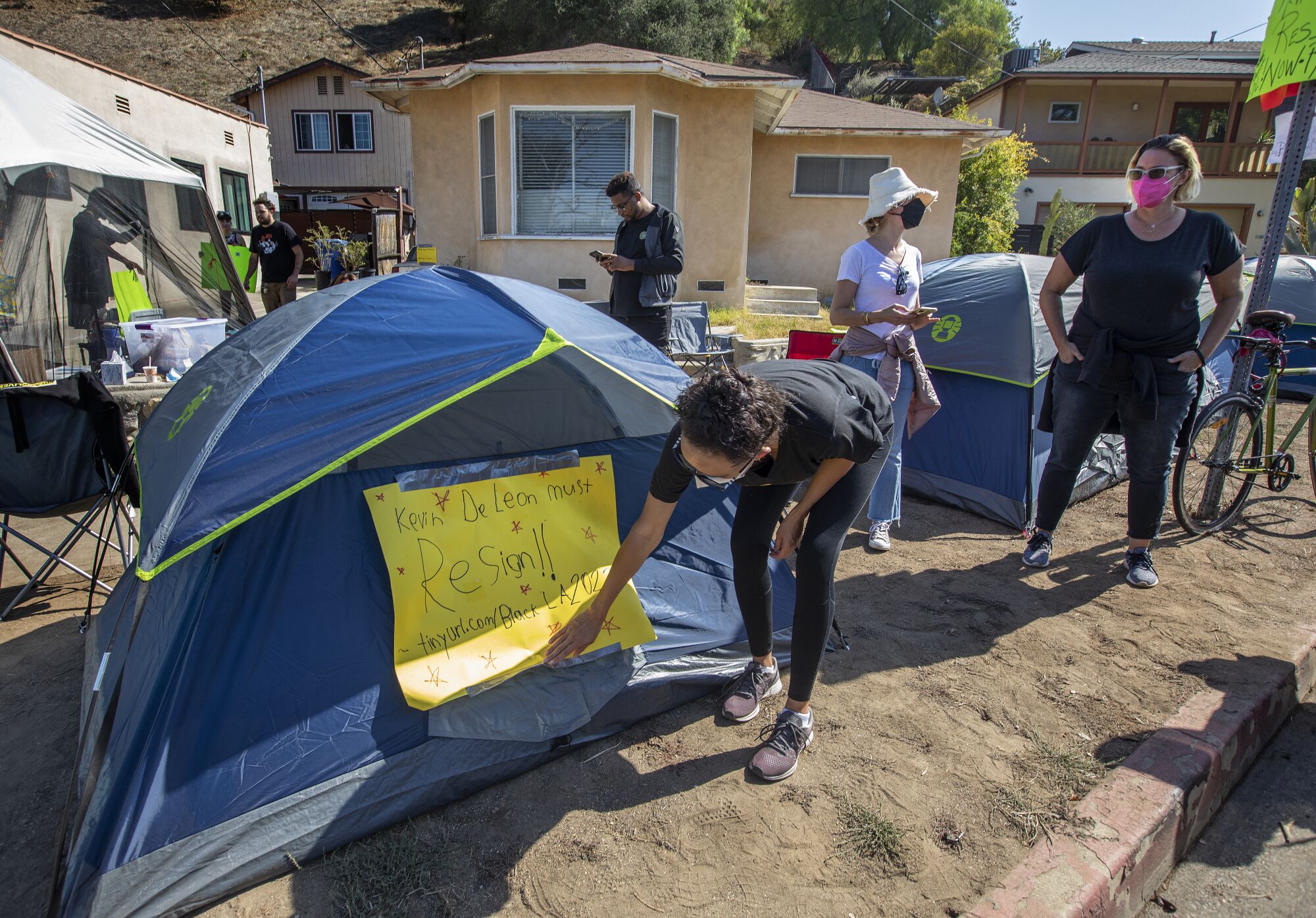 An activist adjusts a sign on one of the tent clusters, reading "Kevin de León must resign!"