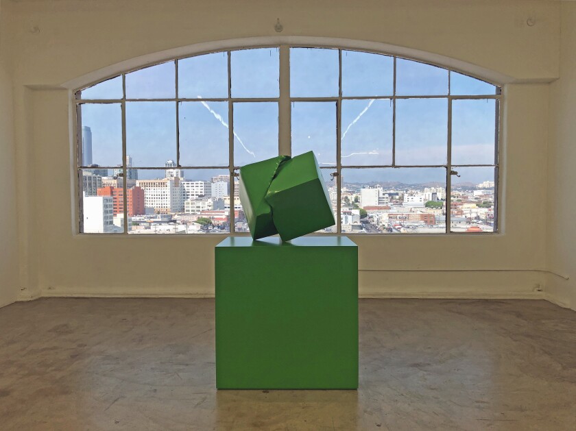 A green geometric sculpture on a green plinth sits before a large window with views of downtown L.A.