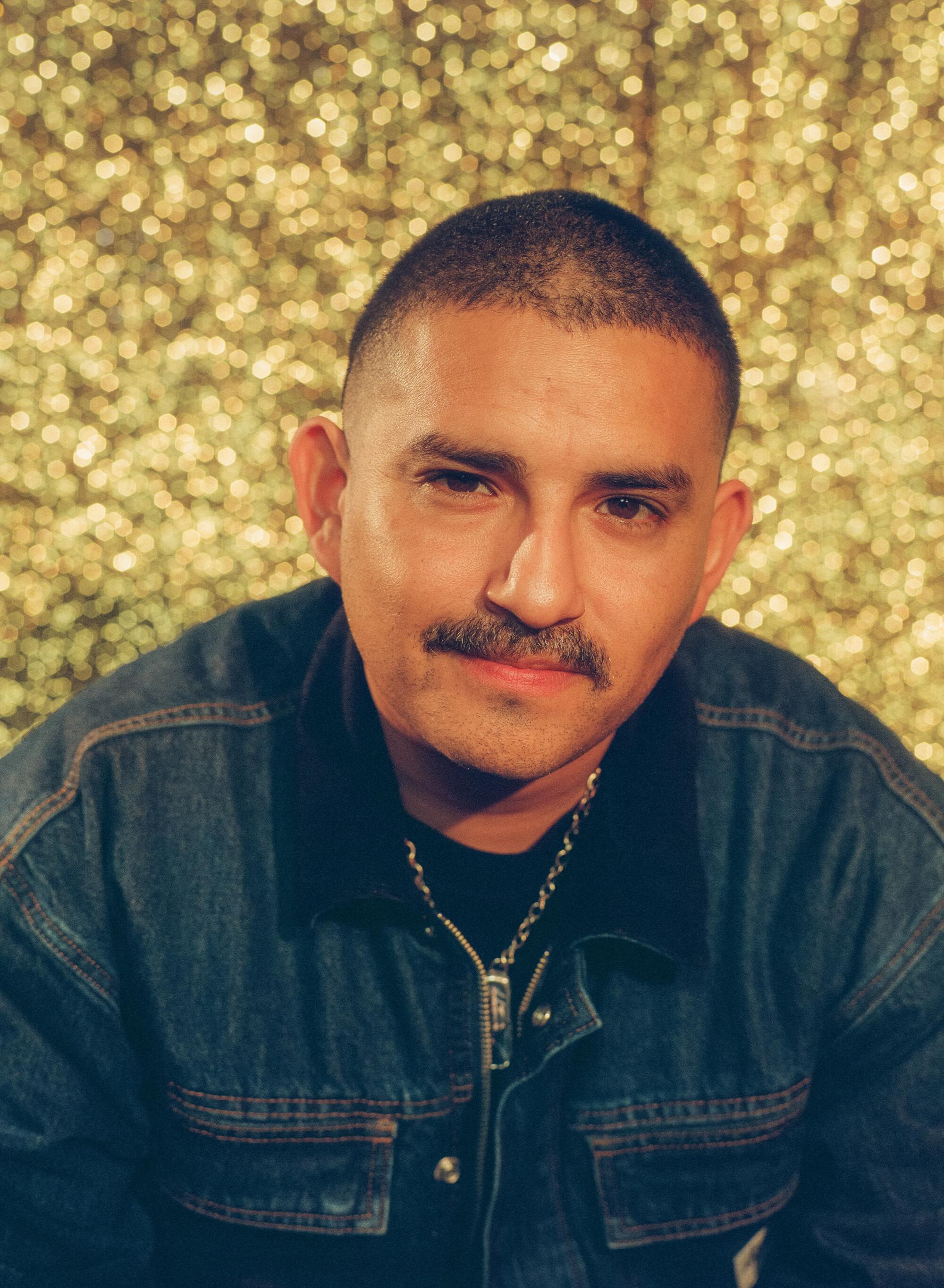 A man in a denim jacket poses in front of a gold glittery background.