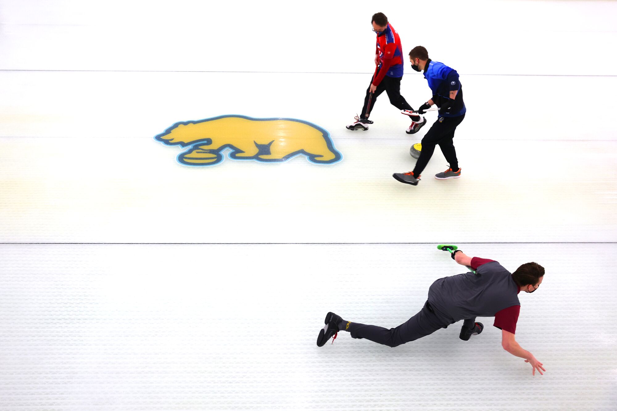 Austin Berndt slides along the ice after throwing a stone as Ken Dethloff, top, and Peter Johns sweep on an adjacent sheet.