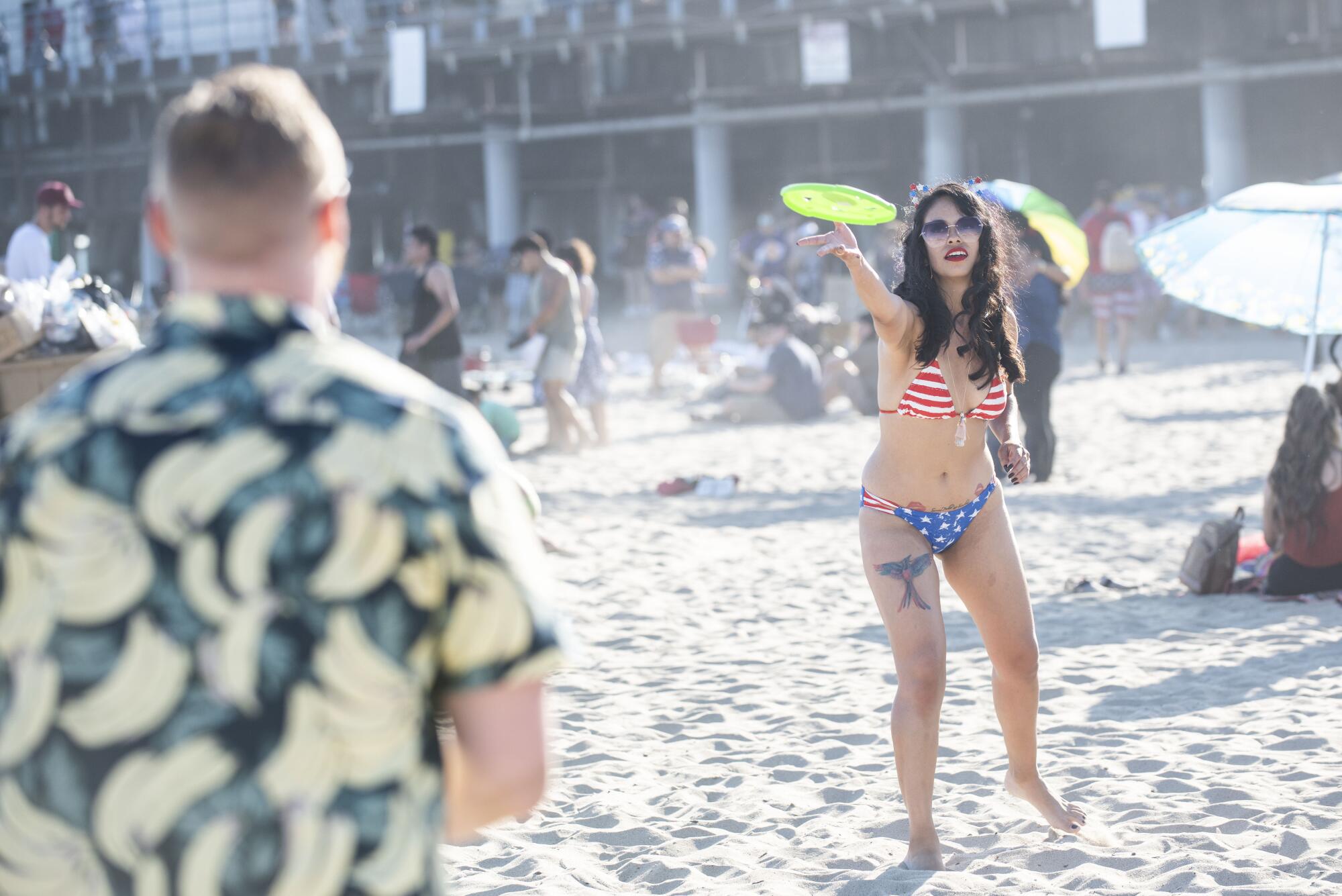 A woman in a red, white and blue bikini throws a green frisbee to a man in a colorful shirt on the beach