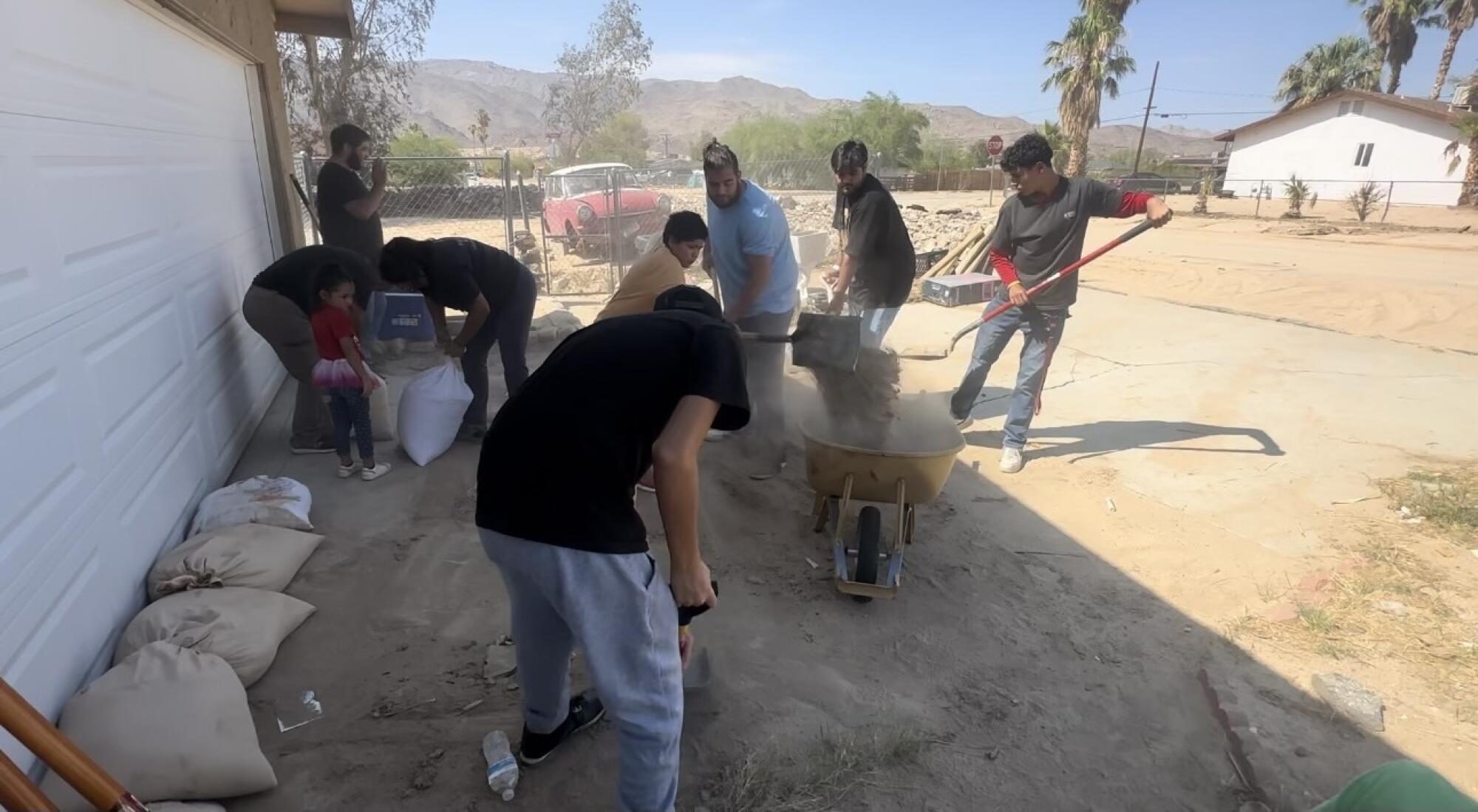 People shovel mud into a wheelbarrow and place sandbags in a driveway.