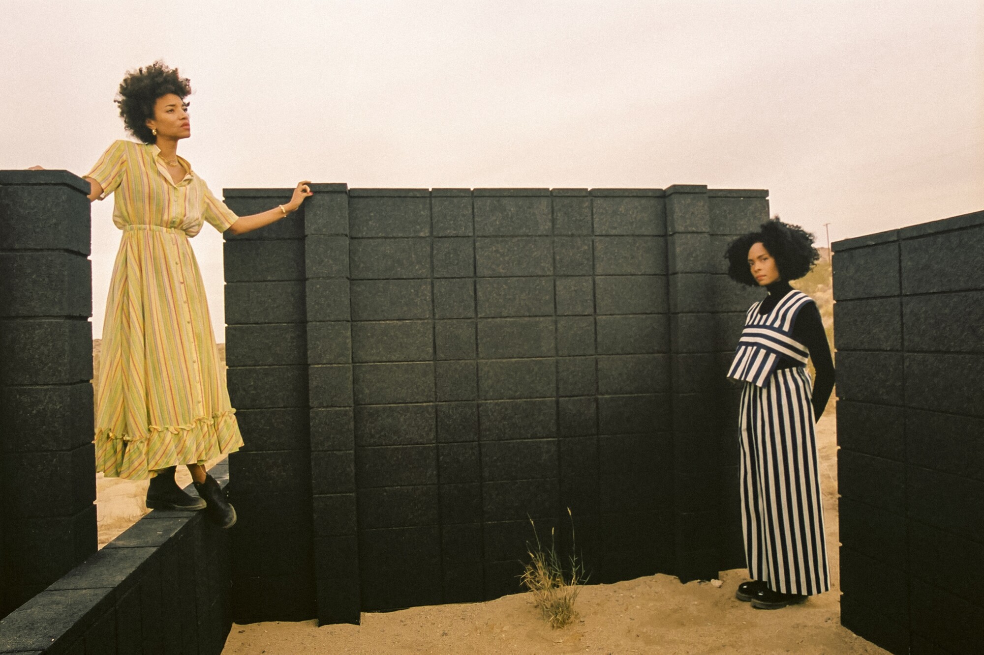 Two women stand amid black cinderblock walls in the desert.