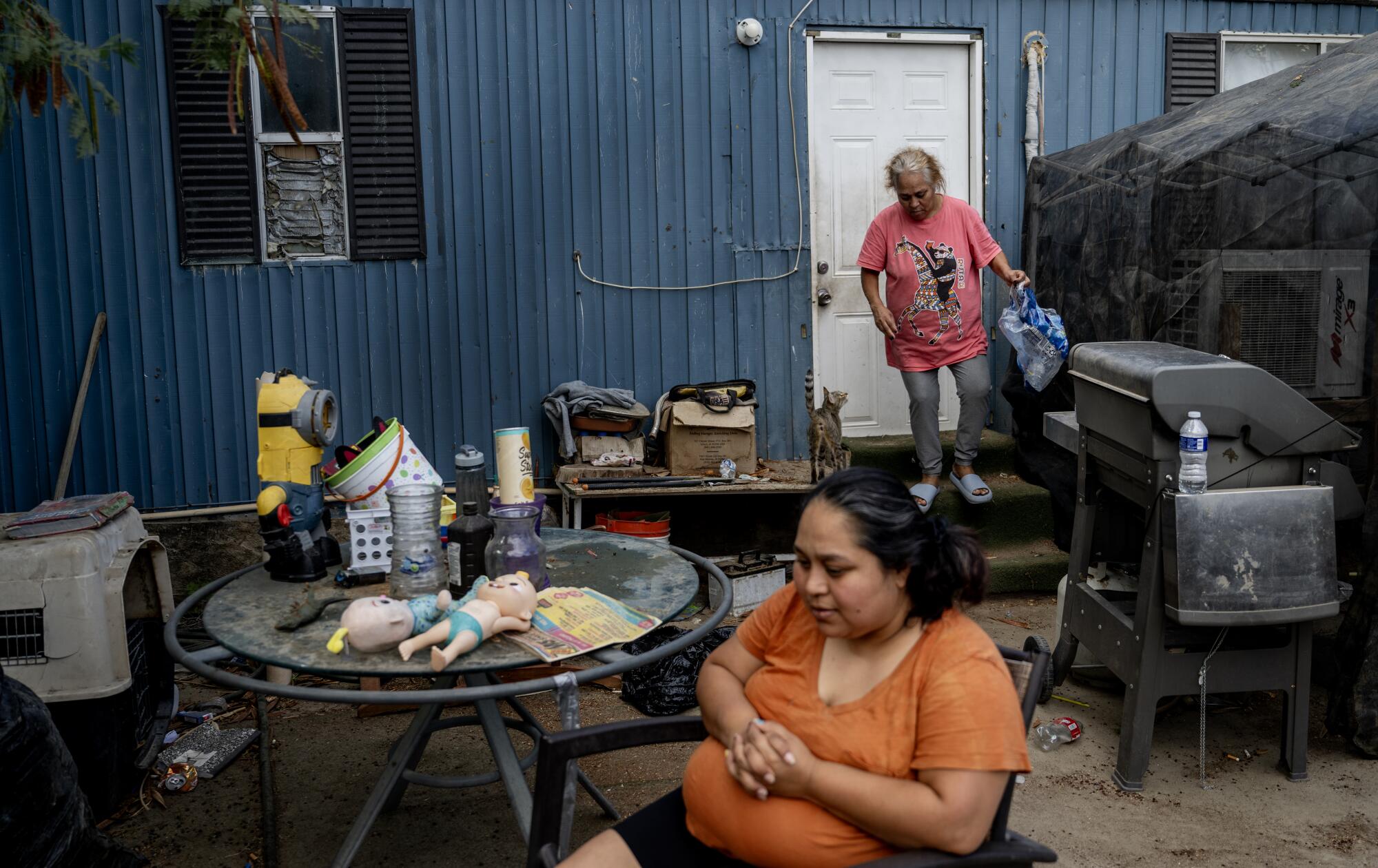 A woman carries bottled water outside her trailer while another woman sits at a table. 